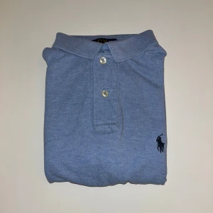 Blå pikétröja Polo Ralph Lauren M - Snygg ljusblå pikétröja från Polo Ralph Lauren i custom slim fit. Klassisk krage, två knappar och broderad logga på bröstet. Tillverkad i mjuk bomull, perfekt för en clean och stilren look.