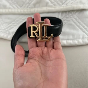 Svart skärp från Ralph Lauren - Stilrent svart skärp från Ralph Lauren med bred rem i skinn och en stor, guldig RL-logga som spänne. Perfekt accessoar för att lyfta din outfit med en lyxig touch och klassisk design.
