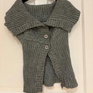 Grå stickad kofta med stora knappar - Superfin grå stickad kofta med korta ärmar och bred krage. Koftan har snygga kabelstickade detaljer och tre stora knappar framtill. Perfekt att slänga över en topp för en chill och trendig look.