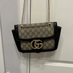 GG axelväska med kedja - Snygg axelväska från Gucci i beige och svart med klassiskt GG-mönster. Väskan har guldfärgad kedjerem och stort GG-spänne framtill. Tillverkad i canvas med detaljer i skinn. Perfekt storlek för det viktigaste och ger en lyxig touch till din outfit.