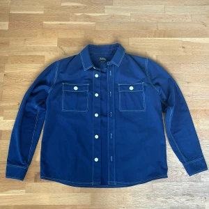 Mörkblå overshirt från A.P.C. - Snygg mörkblå overshirt från A.P.C. med vita kontrastsömmar och stora vita knappar. Skjortan har två bröstfickor med lock och lång ärm. Den är sparsamt använd och i mycket bra skick! Perfekt nu till sommaren!☀️
