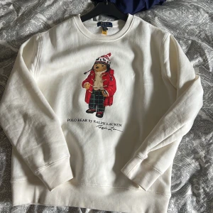 Vit Polo Bear tröja från Ralph Lauren - Vit långärmad sweatshirt från Ralph Lauren med ikoniskt Polo Bear-tryck framtill. Tröjan har rund halsringning och är gjord i mjuk bomull, perfekt för dig som gillar klassisk men lekfull stil. Snygg detalj med björnen i vinteroutfit.