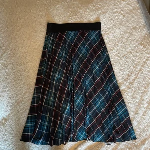 Rutig långkjol från Coccinella - Snygg långkjol från Coccinella med rutigt mönster i blått, svart, vitt och röda detaljer. Kjolen har ett brett svart midjeband och är lätt plisserad för extra volym. Perfekt för dig som vill sticka ut med en cool och färgstark look.