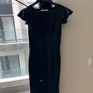Svart satin midi känning från NA-KD - Snygg svart omlottklänning från NA-KD med korta volangärmar och djup v-ringning. Klänningen har spetsdetaljer längs urringningen och midjan samt knytband framtill. Perfekt för dig som gillar stilrena och feminina plagg.