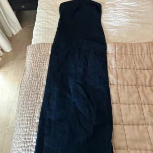 Mörkblå axelbandslös jeansklänning - Snygg långklänning i mörkblå denim med axelbandslös design. Klänningen har en rak och figurnära passform som ger en cool och stilren look. Perfekt för dig som gillar minimalistisk stil och vill sticka ut med denim.