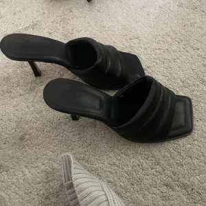 Svarta klackade sandaler med fyrkantig tå - Svarta sandaler från H&M med vadderad rem och fyrkantig tå. Skorna har en smal klack och öppen design, vilket ger en trendig och stilren look. Tillverkade i syntetmaterial och passar perfekt till dig som gillar minimalistisk stil.