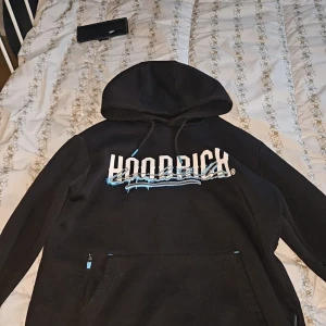 Svart hoodie från Hoodrich med tryck - Svart hoodie från Hoodrich i storlek S med stor vit och blå logga framtill och text på ryggen. Hoodien har huva med snörning, känguruficka och coola detaljer i blått. Perfekt för dig som gillar streetwear och vill sticka ut med snyggt tryck.