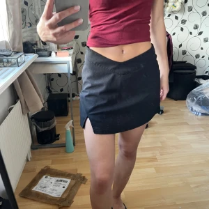 Svart minikjol från Shein med slits - Svart kortkjol från Shein i storlek XS. Kjolen har en snygg v-formad midja och en liten slits framtill. Enkel och stilren design som är lätt att matcha med olika toppar. Midjehöjden går att justeras. Inbyggda shorts under kjolen.