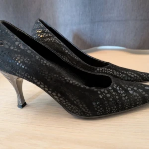 Svarta pumps med mönster från Fraukes - Snygga svarta pumps med spetsig tå och låg klack. Skorna har ett glansigt, reptilinspirerat mönster och är tillverkade i skinn. Perfekta för dig som vill sticka ut med en elegant och unik look.