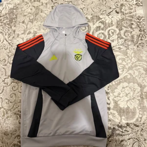 Grå Adidas Benfica hoodie med huva - Snygg grå hoodie från Adidas med Benfica-logga på bröstet, svarta ärmar och röda ränder på axlarna. Tröjan har huva med snörning, half zip och är tillverkad i polyester. Perfekt för fotboll eller chill. Luftig AEROREADY-modell.