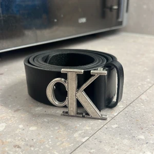 Svart skärp från Calvin Klein - Snyggt svart skärp från Calvin Klein i äkta skinn med en stor, silverfärgad CK-logga som spänne. Bältet har en stilren och klassisk design som passar till jeans eller kostymbyxor. Tillverkat i Rumänien.