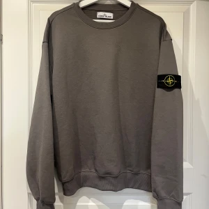 Stone Island - Grå sweatshirt  - Säljer en grå sweatshirt från Stone Island i storlek M. Tröjan har klassisk rund hals, lång ärm och den ikoniska loggan på vänster ärm. Tillverkad i mjuk bomull för skön komfort och stilren look. Perfekt för dig som gillar streetwear och enkel design. Små skador förekommer som på själva serienumret som blivit bränn skadat, men annars är den i ett riktigt fint skick. Fick denna som present, och därav inget kvitto. Vid snabb affär, går priset att diskutera. Några som helst frågor - hör av er! 