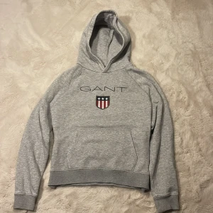 Grå hoodie från GANT  - Snygg ljusgrå hoodie från GANT med broderad logga och sköld på bröstet. Tröjan har huva, magficka och långa ärmar. Perfekt för dig som gillar klassisk och sportig stil. Tillverkad i mjuk bomullsmix för extra komfort.