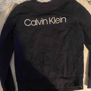 Svart Calvin Klein sweatshirt S - Svart sweatshirt från Calvin Klein i storlek S. Tröjan har rund halsringning, långa ärmar och stor vit logga framtill. Mjuk och skön bomullskänsla, perfekt till jeans eller joggers. Klassisk och stilren design som funkar året runt.