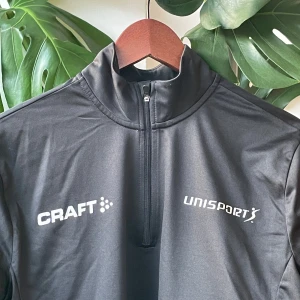 Svart träningströja från Craft - Svart långärmad träningströja från Craft med Unisport-tryck på bröstet. Tröjan har en hög krage och half zip-dragkedja framtill. Tillverkad i ett lätt och stretchigt syntetmaterial som passar perfekt för sport och aktivitet.