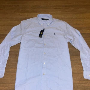 Ljusblå skjorta från Ralph Lauren M - Klassisk ljusblå skjorta från Ralph Lauren med button-down krage och broderad logga på bröstet. Skjortan har långa ärmar, vita knappar och är tillverkad i mjuk bomull. Perfekt för en clean och stilren look.