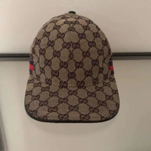 Gucci keps med GG-mönster - Snygg keps från Gucci med klassiskt GG-monogram i mörkbrunt på beige botten. Kepsen har en böjd skärm och ikoniska röd-blå ränder på sidan. Tillverkad i Italien och gjord i slitstarkt bomullsmaterial. Perfekt för dig som vill sticka ut med lyxig streetstyle.