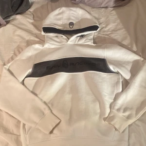Vit hoodie från Sail Racing - Vit hoodie från Sail Racing med svart logga och text på bröstet. Tröjan har huva med svart kant och är tillverkad i en mjuk bomulls- och polyestermix. Perfekt för dig som gillar sportig och clean stil. Pris kan diskuteras vid snabb affär!