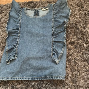 Jeansblus med volangdetaljer - ALDRIG ANVÄND!! Säljer en snygg ärmlös jeansblus i blå denim med volanger längs axlarna och framsidan. Toppen har rund halsringning och en liten öppning med knapp i nacken. Perfekt för dig som gillar en cool och trendig look med extra detaljer. Snyggare på❤️