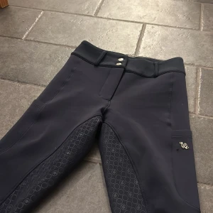Marinblå ridbyxor från zaczess - Snygga marinblå ridbyxor från zaczess i bra skick, de har silikongrepp i sätet och längs benen. Byxorna har stretchigt material, bälteshällor, dubbla knappar i midjan och diskret logga i silver på benet som tyvärr har släppt i ena tråden men det är inget som syns. Även små märken precis vid knäna