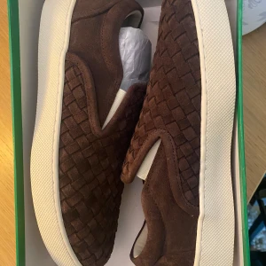 Bruna slip-on sneakers från Bottega Veneta - Snygga slip-on sneakers från Bottega Veneta i brun mocka med flätad design. Skorna har en vit, platt sula och klassisk rund tå. Perfekt för sommar och höst!