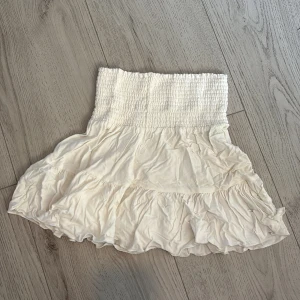 Vit volangkjol med smock - Supersöt vit kortkjol med bred smock i midjan och volangkant. Kjolen är luftig och har en romantisk känsla, perfekt för varma dagar. Den är tillverkad i ett mjukt och lätt tyg som ger en flowy look.