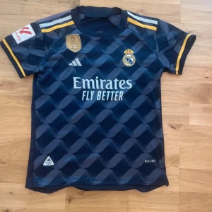 Real Madrid Bellingham tröja Adidas EJ ÄKTA - ⚠️KÖPT I SPANIEN SÅ EJ ÄKTA ⚠️Snygg Real Madrid fotbollströja från Adidas med Bellingham och nummer 5 på ryggen. Tröjan är marinblå med gula detaljer och har ett vågigt mönster. Korta ärmar, slim fit och tillverkad i lätt funktionsmaterial. Perfekt för dig som älskar fotboll och vill sticka ut på planen.