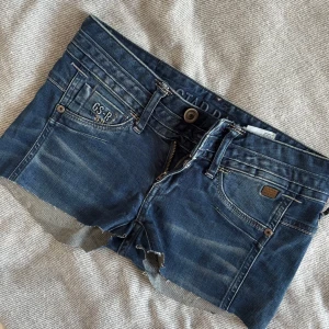 G-Star Raw jeansshorts med rå kant. Lågmidjade! - Snygga blå jeansshorts från G-Star Raw med rå, avklippt kant och klassiska fem fickor. Shortsen har broderad logga, detaljerade bakfickor och slitningar för en cool, avslappnad look. Perfekta för sommardagar när du vill ha en streetig vibe. Jag har klippt de själv, från början var det ett par skinny jeans. OBS båda fickorna där bak har hål i sig!