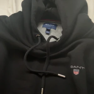 Svart hoodie från GANT med broderad logo - Svart hoodie från GANT med broderad logo och sköld på bröstet. Klassisk huva med snörning och långa ärmar. Mjuk insida och ribbade muddar. Perfekt för chill dagar eller när du vill ha en clean och stilren look.