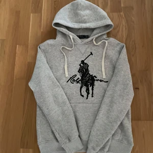Grå hoodie från Polo Ralph Lauren - Snygg grå hoodie från Polo Ralph Lauren med stor svart logga och polospelare på bröstet. Tröjan har huva med vita snören och är tillverkad i mjuk bomullsmix. Perfekt för chill dagar eller när du vill ha en clean streetstyle.