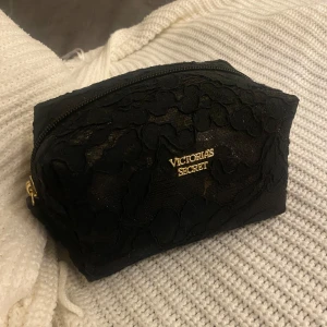 Svart necessär från Victoria's Secret - Snygg svart necessär från Victoria's Secret med blommig spetsdetalj och guldfärgad logga framtill. Perfekt för smink eller småprylar, med dragkedja upptill och kompakt form som är lätt att ta med i väskan.