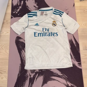 Real Madrid Ronaldo 7 Adidas tröja XS-S - Real Madrid matchtröja i vitt med blå detaljer från Adidas, Climacool-material. Tryck med 'Fly Emirates' och klubbmärke fram, Ronaldo 7 på ryggen. Tre blå ränder på axlarna och broderad jubileumspatch nertill. Perfekt för fans av Ronaldo och Real.