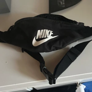 Svart Nike magväska med dragkedja - Säljer en svart magväska från Nike med klassisk vit logga framtill. Väskan har justerbart band och smidig dragkedja upptill. Perfekt för att bära mobilen, plånboken och andra smågrejer när du är på språng.