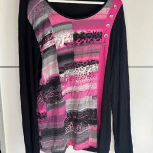 Mönstrad långärmad tröja Brandtex XL - Cool långärmad tröja från Brandtex i svart med färgstarka rosa och grå mönster framtill. Snygga knappdetaljer på axeln och mix av leopard- och randigt print. Mjuk och skön bomullskänsla, perfekt för dig som gillar att sticka ut.