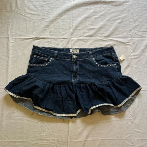 Mörk jeanskjol med volang och detaljer - Midjemått rakt över: 49 cm, längd: 36 cm Supersöt mörkblå jeanskjol från Miss Sixty med volangkant och silvriga detaljer. Kjolen har nitar vid fickorna fram och broderade hjärtan med pärlor på bakfickorna. Perfekt för dig som vill sticka ut med en unik och lekfull stil.