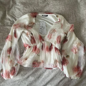 Blommig peplumblus från Gina Tricot - Superfin blommig peplumblus från Gina Tricot i transparent, glansigt material. Blusen är vit med rosa och röda blommor, har v-ringning och långa puffiga ärmar. säljer eftersom att den aldrig kommit till användning, och alltid slutsåld på hemsidan 💕