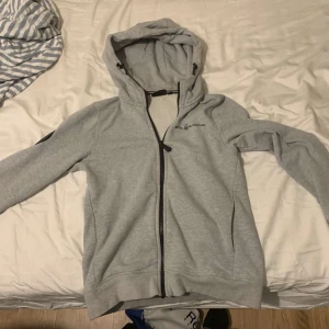 Grå hoodiejacka från Sail Racing - Grå  hoodiejacka från Sail Racing med svart dragkedja och logga på bröstet. Stor tryckt text på ryggen och patch på ena ärmen. Jackan har huva med snörning och är tillverkad i mjuk bomullsmix. Perfekt för chill dagar eller när du vill ha en sportig look.