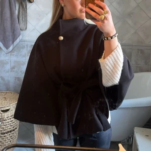 Svart poncho från missäy - Snygg svart kappa med hög krage, dekorativ knapp vid halsen och knytband i midjan. Modellen är vid och har en cape-liknande form som ger en cool och trendig siluett. Perfekt för dig som gillar stilrena och unika plagg.