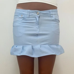 Ljusblå jeanskjol med volangkant - Supersöt ljusblå kort jeanskjol med volang längst ner. Kjolen har klassiska jeansdetaljer som fickor fram och bak, bälteshällor och knappstängning. Perfekt för dig som vill ha en söt och trendig look till sommaren.