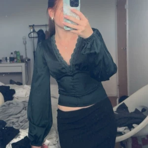 Mörkgrön blus med spets från BikBok - Supersnygg mörkgrön blus från BikBok med v-ringning och spetskant. Modellen är croppad med långa puffärmar och knappar framtill. Perfekt för dig som gillar en feminin och trendig look.