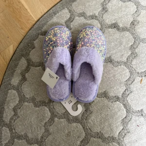 Lila fluffiga slippers med paljetter - Supermysiga slippers i lila med fluffigt syntetmaterial och glittriga paljetter på ovansidan. Perfekta att glida runt i hemma när du vill ha det extra bekvämt och ändå sticka ut lite. Sula i vitt och öppen häl för enkel påtagning.