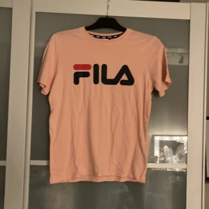 Ljusrosa FILA t-shirt med logga - Snygg ljusrosa t-shirt från FILA med stor logga framtill i svart och rött. Klassisk rund halsringning och korta ärmar. Skön och mjuk bomullskvalitet, perfekt till en avslappnad stil.