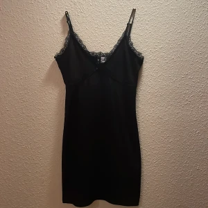 Svart kort klänning med spetsdetaljer - Supersnygg svart kort klänning med tunna axelband och v-ringning. Klänningen har fina spetsdetaljer längs urringningen och över bysten, samt en liten rosett framtill. 