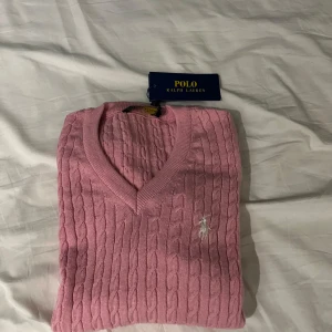 Rosa kabelstickad tröja Polo Ralph Lauren - Supersnygg rosa kabelstickad tröja från Polo Ralph Lauren med v-ringning och broderad logga på bröstet. Tröjan har lång ärm och är gjord i mjuk bomull, perfekt för lager på lager. Klassisk och stilren modell som lyfter din outfit.