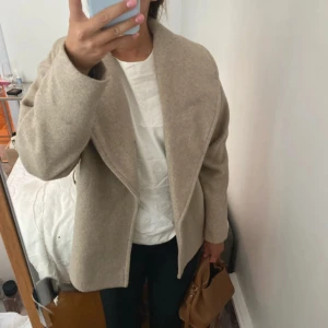 Beige omlottjacka med bälte - Snygg beige omlottjacka med bred krage och bälte i midjan. Jackan har en avslappnad passform och är tillverkad i ett mjukt ullblandat material. Perfekt för lager-på-lager och funkar lika bra till jeans som till klänning.