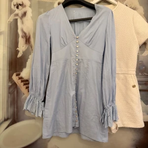 Adoore- Ljusblå kortklänning med volangärm - Supersöt ljusblå kortklänning i 100% bomull med v-ringning, knappar framtill och långa ärmar med volang och broderade detaljer vid ärmslutet. Perfekt för dig som gillar romantiska och feminina vibes. Klippt av lappen bak men från märket Adoore.