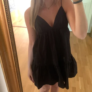 Svart kort klänning från Zara - Supersöt svart kort klänning från Zara med tunna axelband och v-ringning. Klänningen har volang nertill och knytning i ryggen för en snygg detalj. Perfekt för varma dagar och enkel att styla med smycken eller sneakers. Borttagen lapp men bara testats. 