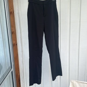 Svarta kostymbyxor med raka ben - Snygga svarta kostymbyxor med klassisk rak passform och hällor för bälte. Byxorna har diskreta sidfickor och stängs framtill. Perfekta för en clean och stilren look. Materialet känns mjukt och följsamt.