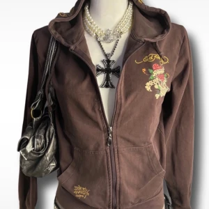 Brun Ed Hardy hoodie med döskalletryck - Säljer min as coola ed Hardy hoodie då den tyvärr inte kommer till användning nå mer! Köpt på vinted. Defekter: ett litet hål i armhålan (märks inte när man har på sig) några kristaller har ramlat av på döskallen. (Vissa lånade bilder från förra ägaren) storlek m men passar s och xs!