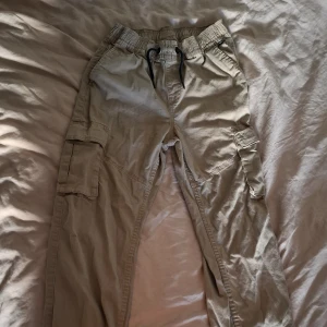 Beige cargobyxor från Kappahl, 164 - Snygga beige cargobyxor från Kappahl i storlek 164. Byxorna har elastisk midja med snörning och flera praktiska fickor på sidorna. Tillverkade i mjuk bomull för en skön känsla och avslappnad look.